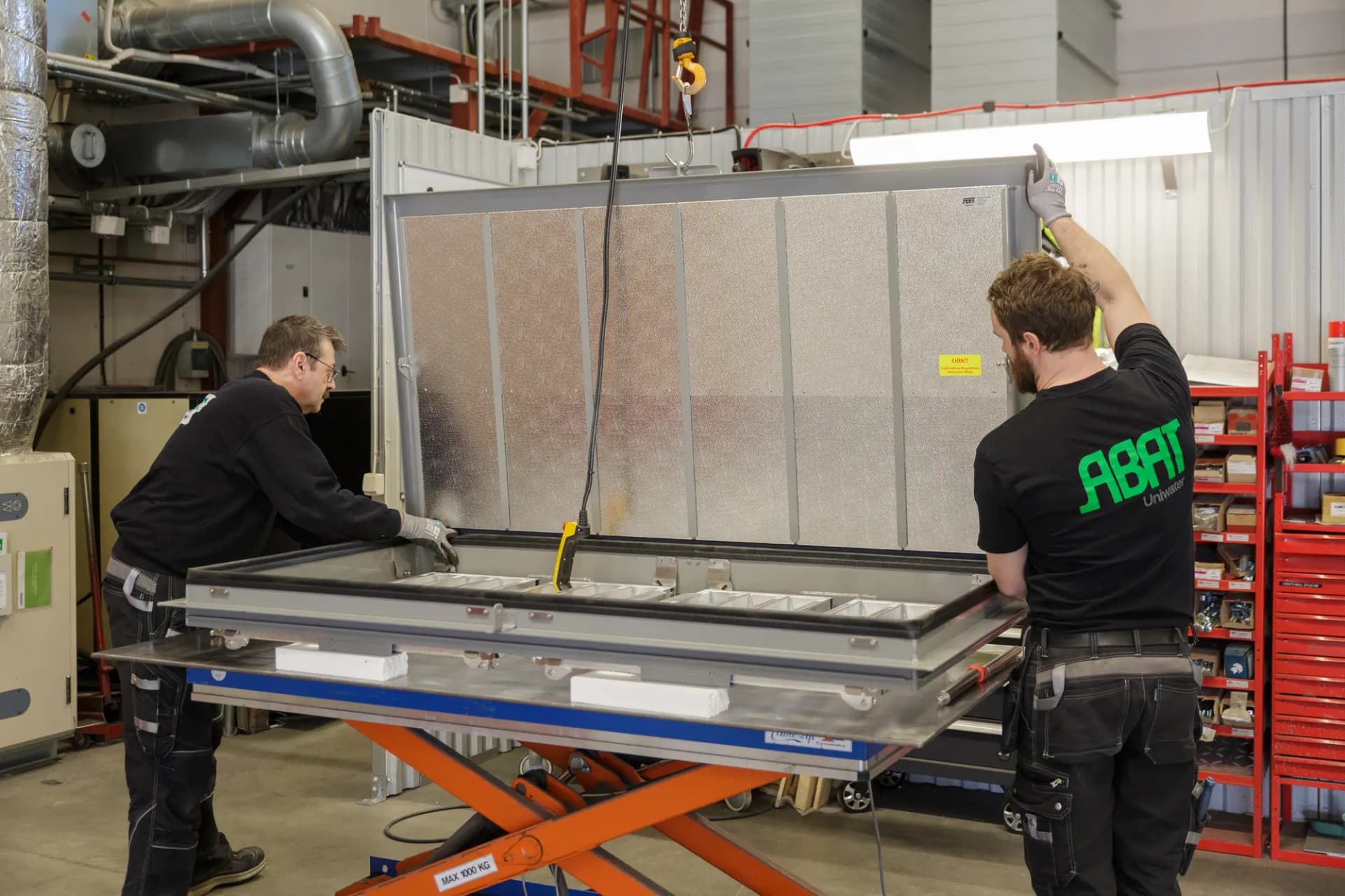 Två medarbetare lyfter en stor aluminiumlucka på ett lyftbord i ABAT:s verkstad